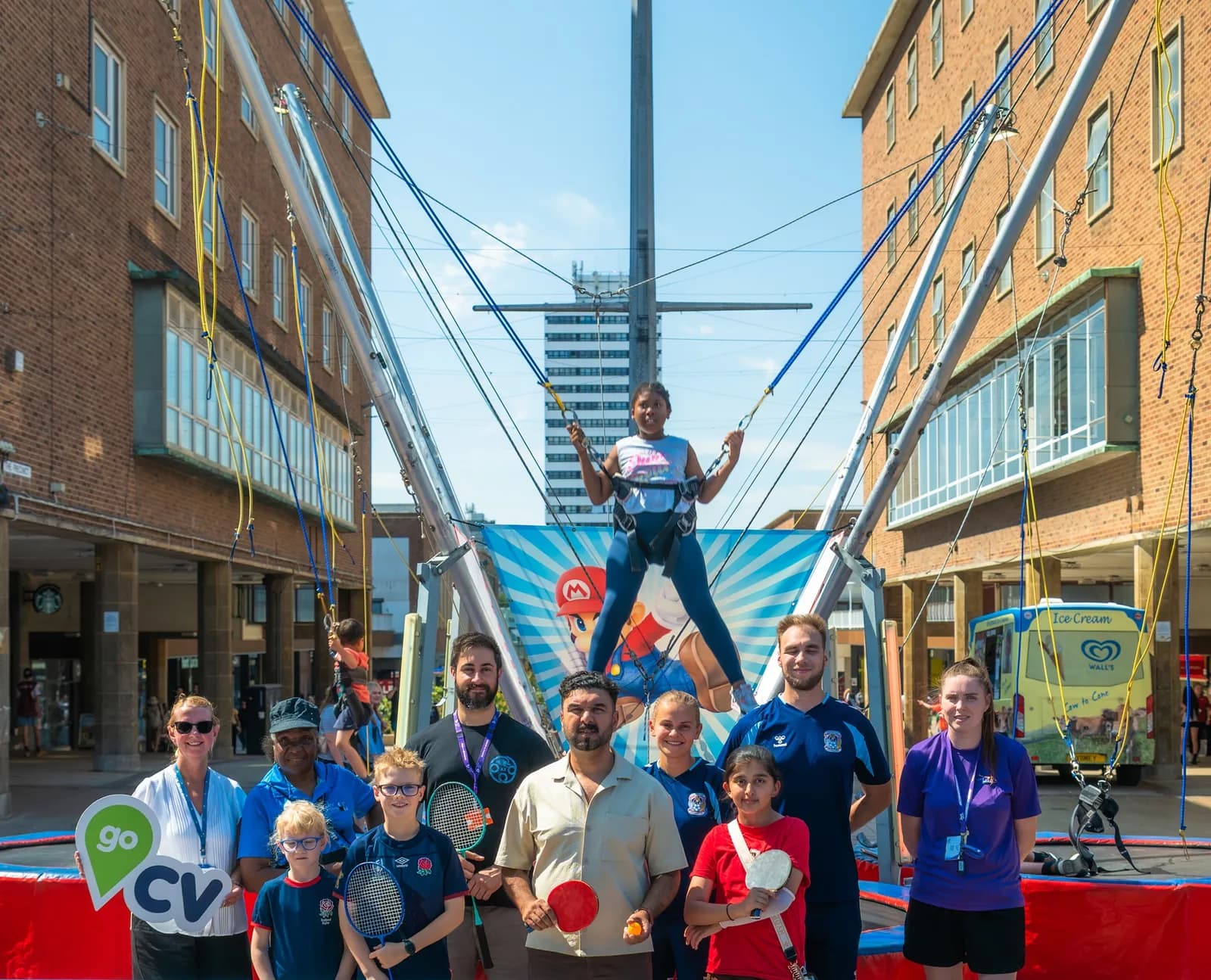 A group of adults and children stand in front of a trampoline bungee jump, where one child is suspended in the air, with a city street as the background.