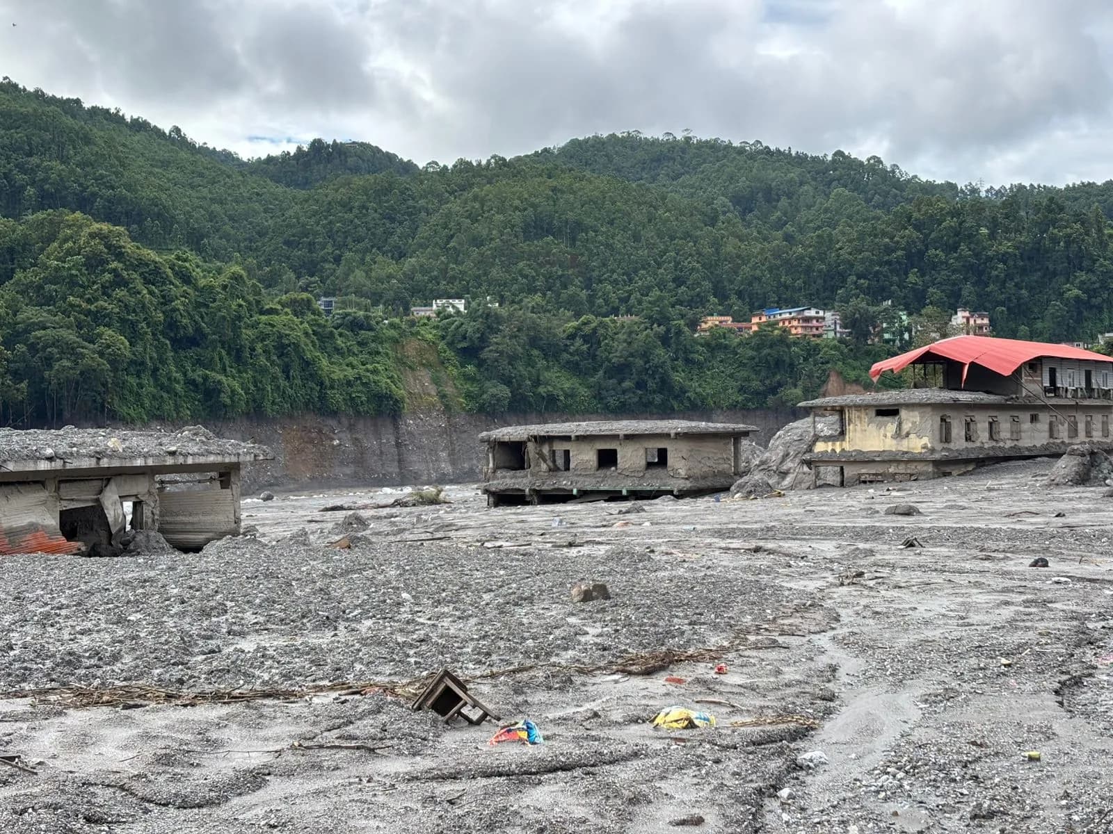 Three damaged concrete buildings sit on a desolate, debris-filled landscape with green, tree-covered hills and a cloudy sky in the background.