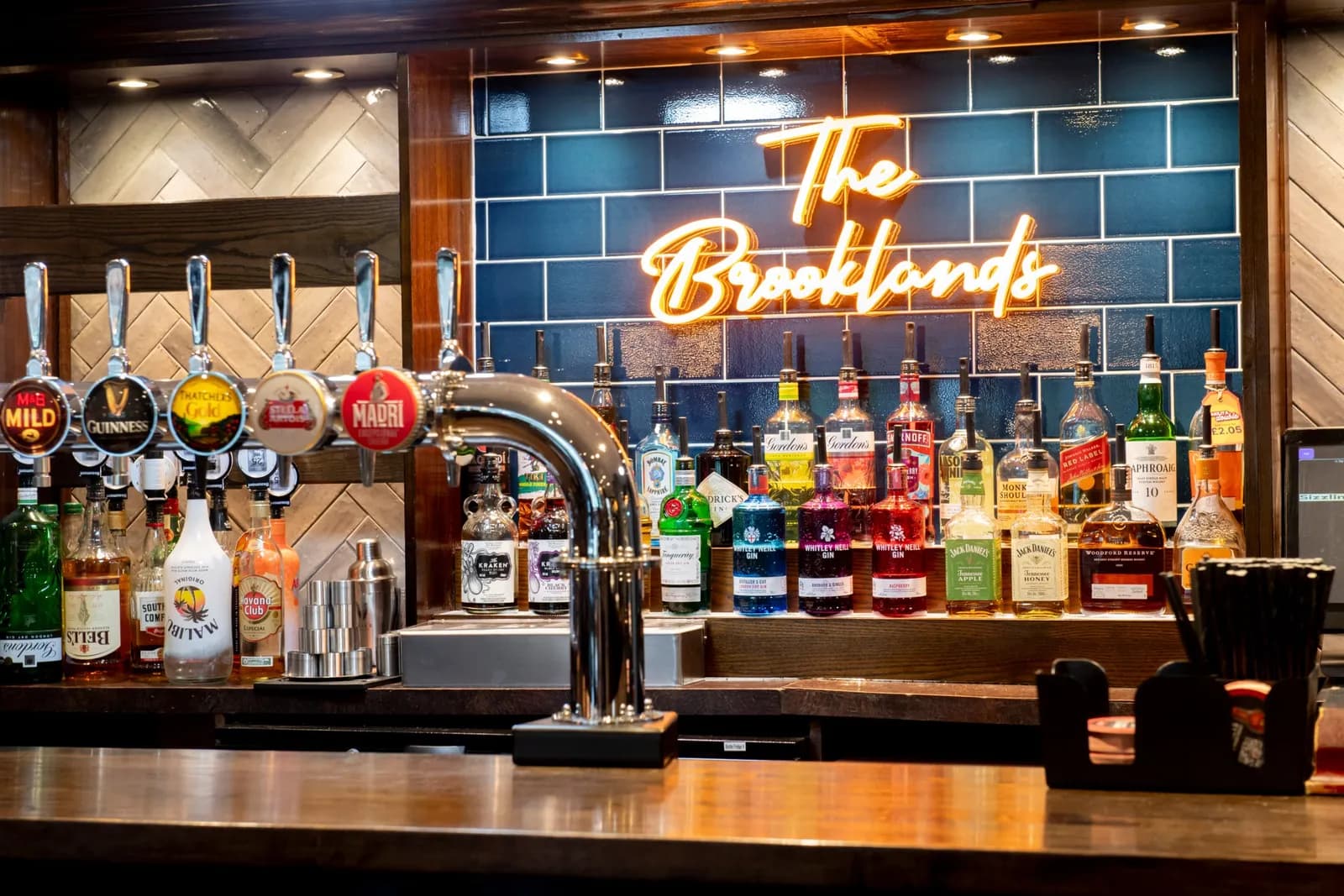 A well-stocked bar features several beer taps, various liquor bottles, and a neon sign glowing "The Brooklands" against a tiled wall.